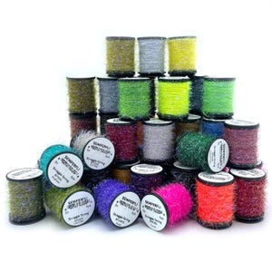May include: A collection of colorful spools of Semperfli Straggle String, in shades of green, yellow, pink, purple, and silver. The spools are cylindrical and stacked, with labels visible on some. Ideal for crafting and fly tying.