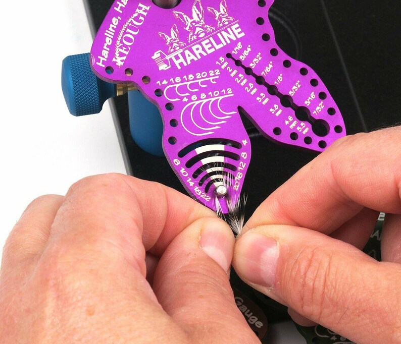 May include: A purple Hareline Keough fly tying gauge tool with various measurement markings. The tool is being used to measure a small bundle of fly tying material. The tool has the text "Hareline" and "Keough" printed on it.
