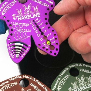 May include: A purple Hareline hook and bead gauge with a blue knob, featuring measurements in inches and fractions of an inch. The gauge has multiple holes and markings for precise measurements. The text "HARELINE" and "HARELINE.COM" are visible.