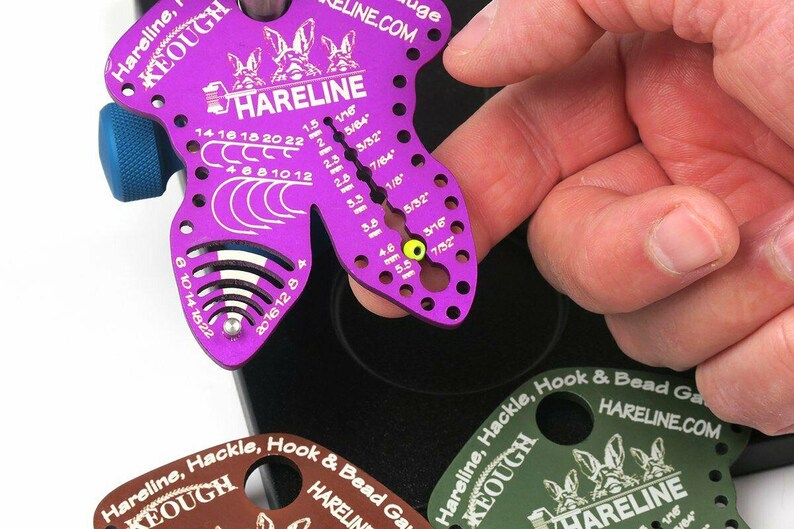 May include: A purple Hareline hook and bead gauge with a blue knob, featuring measurements in inches and fractions of an inch. The gauge has multiple holes and markings for precise measurements. The text "HARELINE" and "HARELINE.COM" are visible.