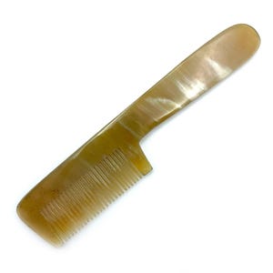 May include: A horn comb with a long handle and fine teeth. The comb is a light brown color with a marbled pattern. The handle is smooth and rounded, while the teeth are evenly spaced. The comb is designed for hair care.
