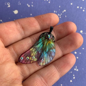 May include: A colorful pendant in the shape of a butterfly wing, featuring iridescent hues of green, purple, and yellow. The wing is adorned with small, sparkling rhinestones and a black metal setting. The pendant is held in a hand.
