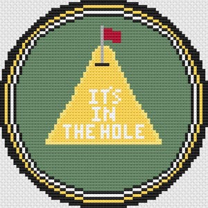 May include: A cross-stitch design featuring a golf-themed emblem. The design includes a yellow triangle with the text "IT'S IN THE HOLE" and a red flag on a pole, set against a green background, all within a circular frame.