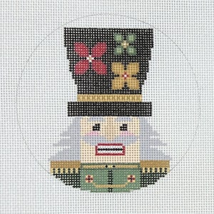 May include: A cross-stitch design of a nutcracker soldier. The soldier wears a black hat with floral accents, a green jacket, and a tan face. The design is circular and set against a white background.