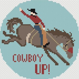 May include: A circular cross-stitch pattern on a light blue background featuring a brown bucking horse and a rider in a red shirt, black hat, and blue jeans. The text COWBOY UP! is stitched in red letters below the horse and rider figure.