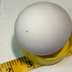 May include: A white egg with a small hole, resting on a yellow egg cup. A yellow measuring tape is in the foreground, with the numbers 2 and 3 visible. The egg cup is approximately 1 inch in diameter.