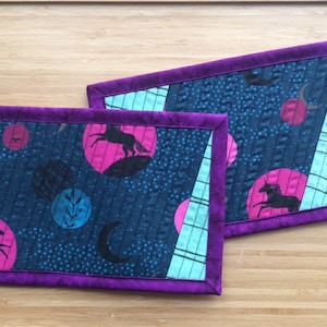 Peut inclure: Deux dessous-de-verre matelassés avec une bordure violette, un fond bleu foncé et un motif de licorne rose et noire. Les dessous-de-verre présentent un motif répétitif de lunes, d'étoiles et de licornes.