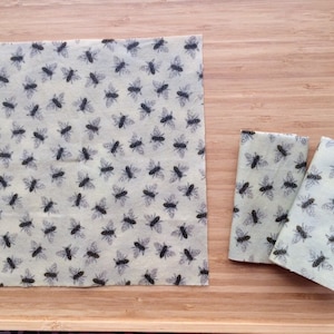 May include: Three pieces of beeswax food wrap with a bee pattern. The wrap is a light yellow colour with black bees printed on it. The wrap is folded and laid out on a wooden surface.
