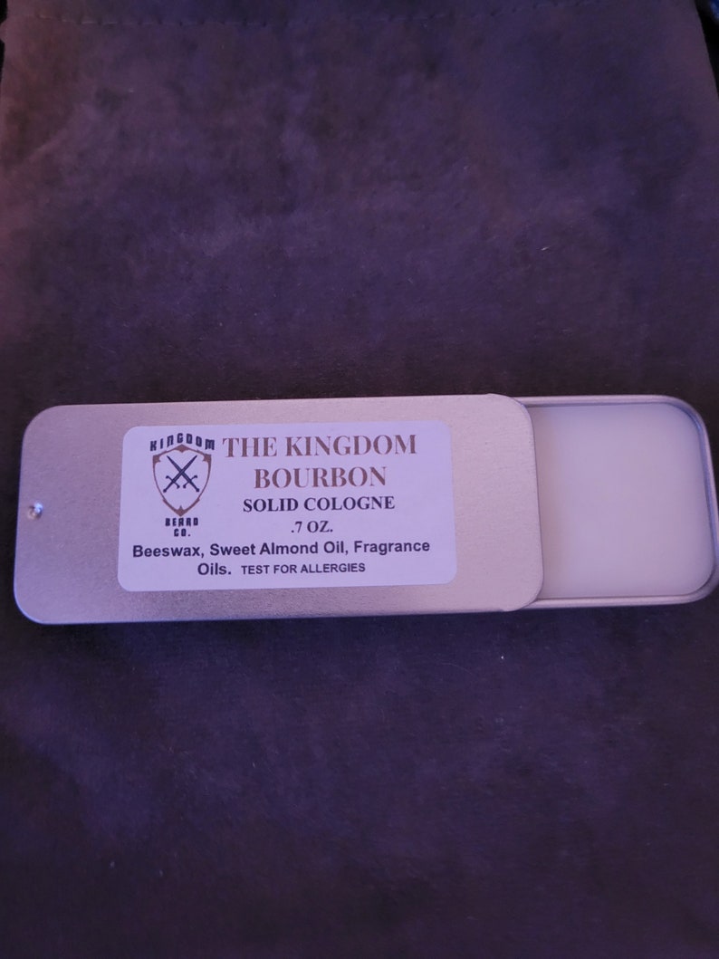 May include: A silver tin with a white label that reads "The Kingdom Beard Co. Bourbon Solid Cologne .7 oz. Beeswax, Sweet Almond Oil, Fragrance Oils. Test for Allergies."