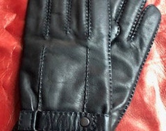 Op de afbeelding: Een paar zwarte leren handschoenen met decoratieve stiksels en een band met een knoopsluiting. De handschoenen worden gepresenteerd op een rode ondergrond.