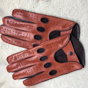 Könnte beinhalten: Ein Paar braune Leder-Fahrerhandschuhe mit perforierten Details und schwarzem Lederbesatz. Die Handschuhe haben einen Druckknopfverschluss am Handgelenk.