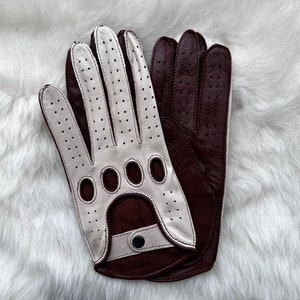 以下が含まれることがあります： 革製の運転用手袋のペア。1つは白地に茶色のアクセント、もう1つは茶地に白のアクセント。手袋は穴あきデザインで、ボタン留めです。