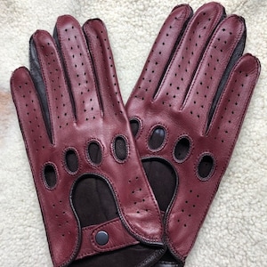 Könnte beinhalten: Ein Paar burgunderfarbene Leder-Fahrerhandschuhe mit perforierten Details und schwarzem Besatz. Die Handschuhe sind fingerlos und haben einen Riemen am Handgelenk.