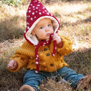 May include: A red and white polka-dot felted hood with a pointed top and tie strings, worn over a mustard yellow chunky knit cardigan featuring a textured bobble pattern, dark buttons, and long sleeves, paired with dark teal trousers and brown shoes.