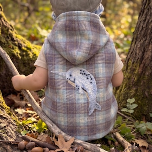 May include: A sleeveless hooded vest featuring a light blue and brown plaid pattern with a light-colored t-shirt underneath. An embroidered narwhal appliqué is centered on the back of the vest. A grey cap is worn on the head.