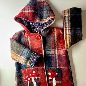 Peut inclure: Veste à capuche rouge et noire à carreaux avec une poche à rabat en forme de champignon. La veste est doublée d'un tissu floral.