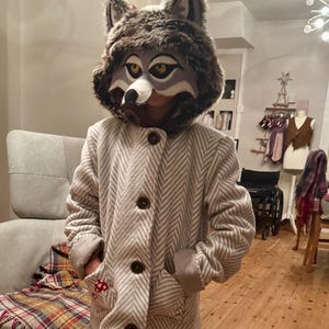 May include: A child wearing a grey and white herringbone jacket with a wolf mask. The mask has a grey and brown fur hood and a white muzzle with black eyes.