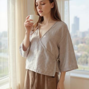 May include: A woman wearing a light grey wrap top with wide sleeves and a brown skirt. The top has a V-neck and ties at the side. The skirt has a drawstring and pockets. She is holding a white cup.