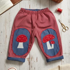 May include: Red and white striped trousers featuring blue denim knee patches with embroidered red and white mushroom designs. The trousers have an elastic waistband and blue denim cuffs at the bottom of each leg, showcasing a detailed mushroom motif.