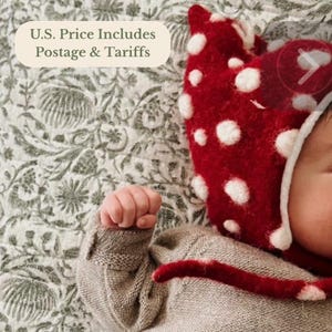 Peut inclure: Un bonnet rouge foncé orné d'un motif à pois blancs et d'un lien de serrage, porté par un enfant vêtu d'un pull en tricot beige clair à manches longues. Le bonnet présente un aspect texturé et duveteux avec un bord blanc.
