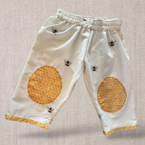 May include: Cream-coloured baby trousers with an elasticated waist, decorated with small black bee motifs. The knees have large, circular honeycomb patches. The cuffs are trimmed with a honeycomb pattern.