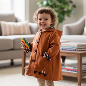 May include: A rust-colored hooded coat featuring a button-front closure, long sleeves with patterned cuffs, and two front pockets. One pocket displays a small mushroom graphic and black triangular accents along the bottom hem of the garment.