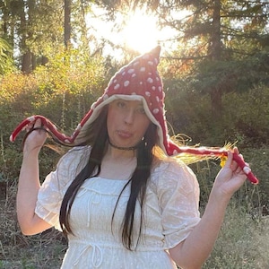 May include: A red pointed hat with white spots resembling a mushroom cap, featuring long side flaps with white spots and a cream-colored inner lining. The hat is made of a textured knit material and has a distinct conical shape at the top.