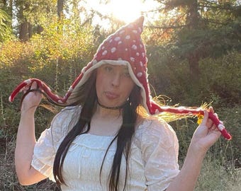 May include: A red pointed hat with white spots resembling a mushroom cap, featuring long side flaps with white spots and a cream-colored inner lining. The hat is made of a textured knit material and has a distinct conical shape at the top.
