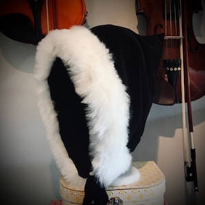 May include: A black hood featuring a thick, plush white faux fur trim along the front opening. The hood is constructed from a solid black fabric and includes black fabric ties at the base of the neck to secure the garment in place when worn.