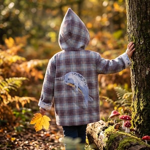 May include: A hooded jacket featuring a blue and brown plaid pattern with a light blue narwhal appliqué on the back. The jacket has long sleeves with folded cuffs and a pointed hood, worn over dark blue pants while holding a yellow maple leaf.