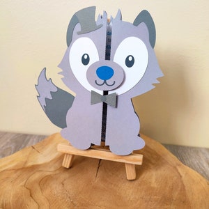Peut inclure: Un loup en papier cartonné gris portant un chapeau haut de forme et une cravate, debout sur un chevalet en bois. Le loup a un nez bleu et de grands yeux blancs.