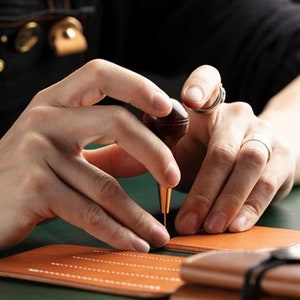 May include: A person using a leather stitching tool to sew a brown leather wallet. The tool has a wooden handle and a metal needle. The leather is being stitched with white thread.