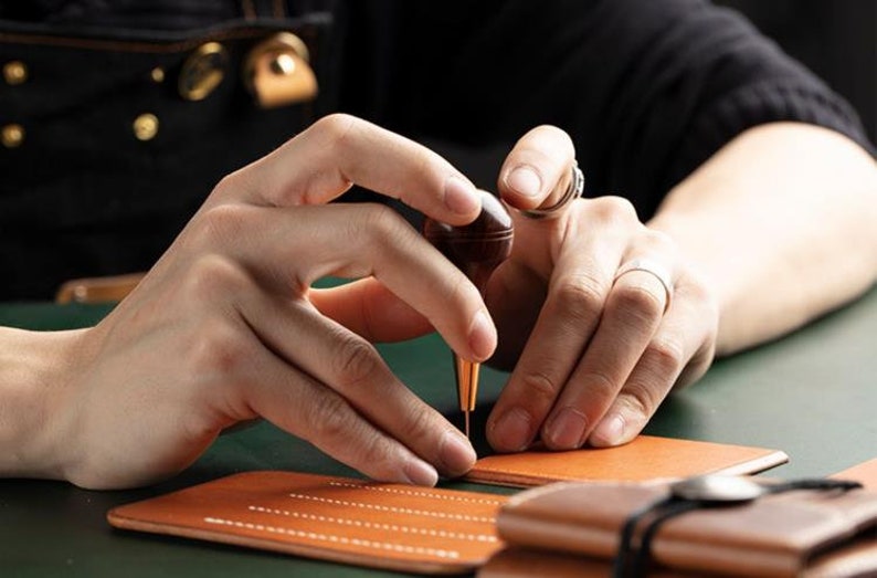 May include: A person using a leather stitching tool to sew a brown leather wallet. The tool has a wooden handle and a metal needle. The leather is being stitched with white thread.