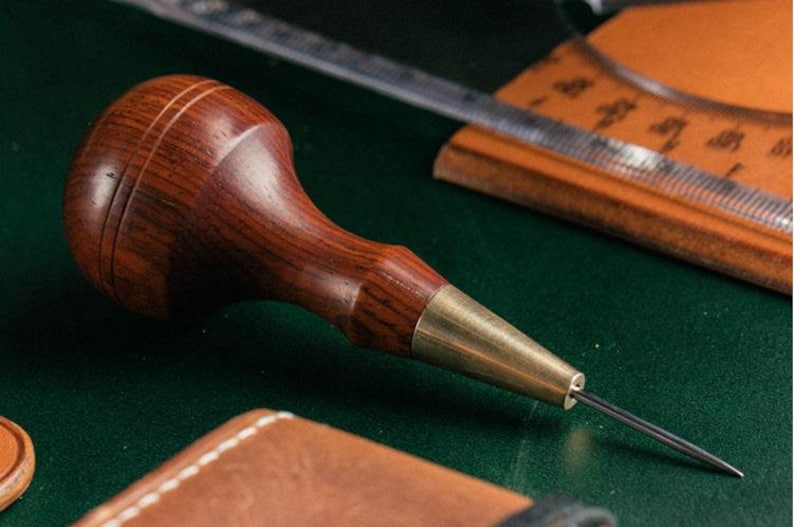 May include: A close-up of a wooden leather stitching awl with a brass tip. The awl is on a green surface.