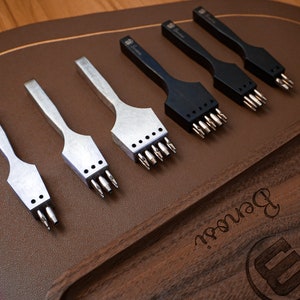 May include: Five leather hole punch tools with different sized punches. The tools are arranged in a row on a brown leather surface. The tools are made of metal and have black and silver handles.