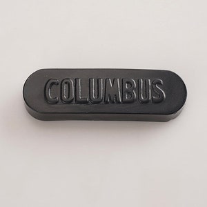 K&ouml;nnte beinhalten: Ein schwarzer rechteckiger Knopf mit dem Wort "COLUMBUS" in wei&szlig;er Pr&auml;gung.