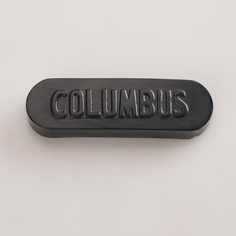 K&ouml;nnte beinhalten: Ein schwarzer rechteckiger Knopf mit dem Wort "COLUMBUS" in wei&szlig;er Pr&auml;gung.