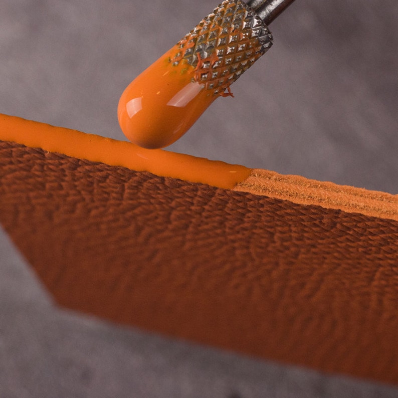 May include: A close-up of a tool applying orange paint to a piece of brown leather. The tool has a metal handle and a small, round tip.