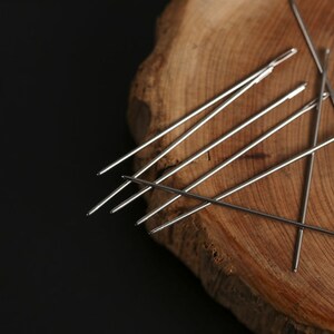 May include: A set of silver sewing needles arranged on a wooden surface. The needles are sharp and pointed, and they are all the same size.