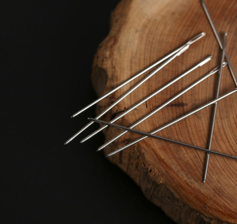 May include: A set of silver sewing needles arranged on a wooden surface. The needles are sharp and pointed, and they are all the same size.