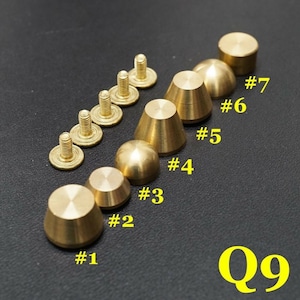 Puede incluir: Nueve tachuelas de metal de color dorado en forma de cono con base de tornillo. Las tachuelas están dispuestas en una fila de la más grande a la más pequeña, con la tachuela más grande en la parte superior de la imagen. Las tachuelas están etiquetadas con los números del 1 al 9.