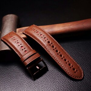 May include: Two brown leather watch bands with red stitching and a perforated design. The bands are lying on a dark brown surface.