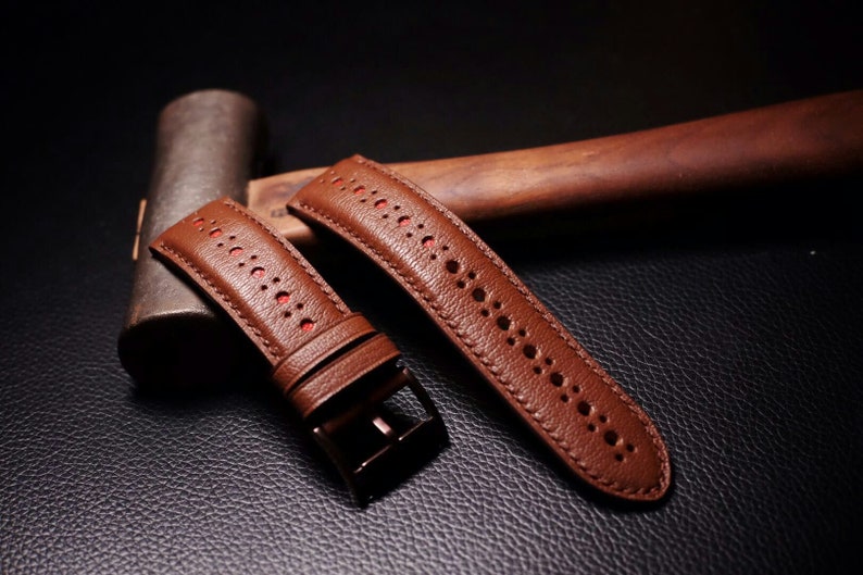 May include: Two brown leather watch bands with red stitching and a perforated design. The bands are lying on a dark brown surface.