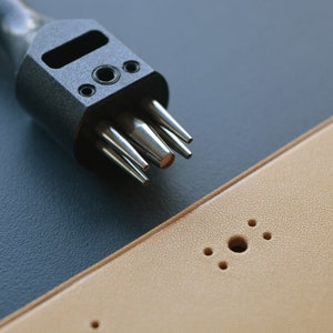 May include: A close-up of a black leather punch tool with multiple punch sizes. The tool is lying on a dark blue surface next to a piece of tan leather with multiple holes punched in it.