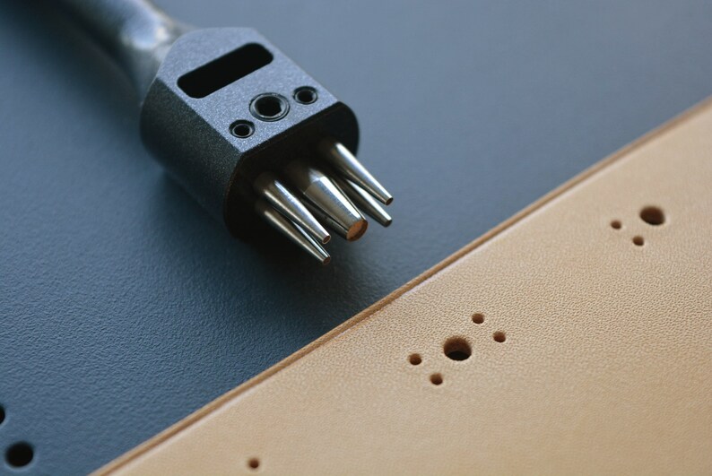 May include: A close-up of a black leather punch tool with multiple punch sizes. The tool is lying on a dark blue surface next to a piece of tan leather with multiple holes punched in it.