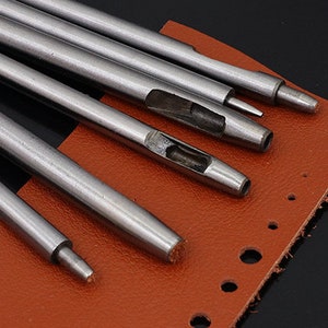 May include: A set of six metal leather hole punches of varying sizes, arranged on a brown leather surface. The punches are used for creating holes in leather for sewing or other crafts.