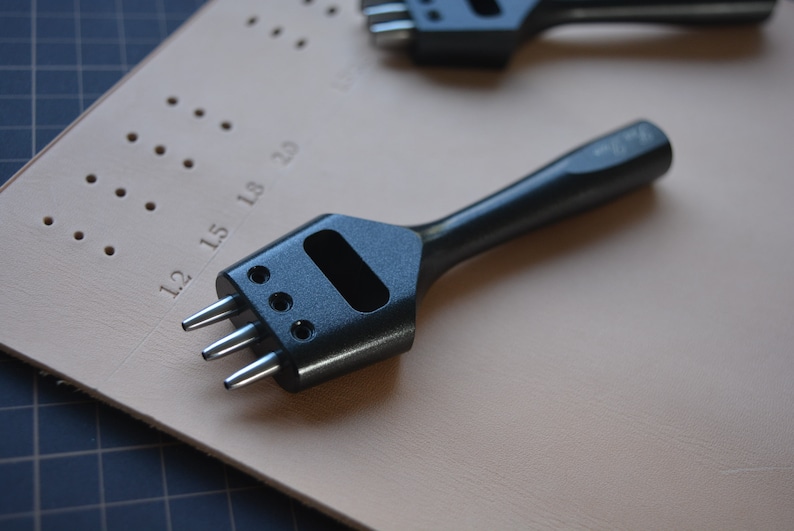 Pu&ograve; includere: Uno strumento di punzonatura in pelle nero con tre punte, utilizzato per fare buchi nella pelle. Lo strumento &egrave; appoggiato su un pezzo di pelle beige con segni numerati.