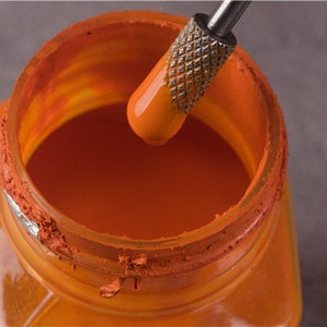May include: A close-up of a glass jar filled with orange paint. A metal tool is being used to stir the paint.