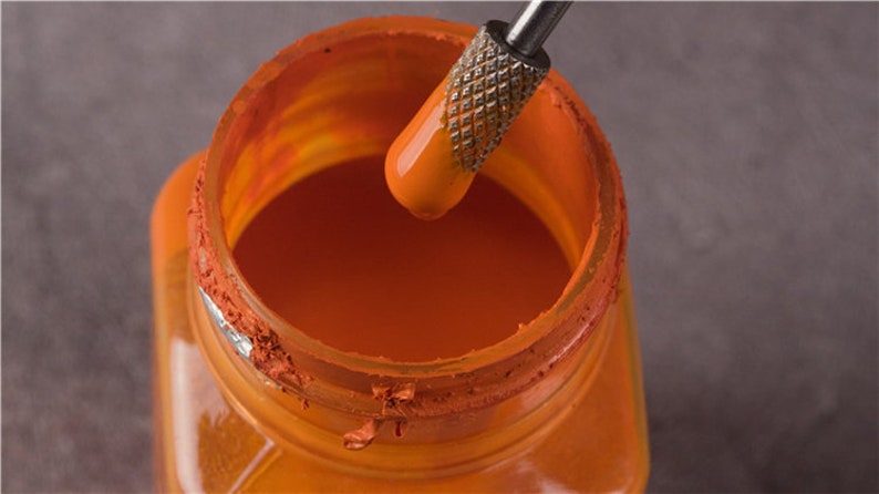 May include: A close-up of a glass jar filled with orange paint. A metal tool is being used to stir the paint.