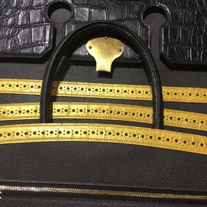 May include: Close-up of a black leather handbag with gold-colored straps. The straps have a repeating pattern of small circles.